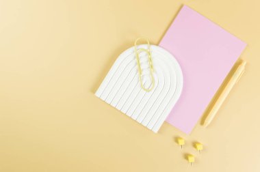 Pembe defterin üst görüntüsü, sarı kalem, ataç, raptiyeler, sarı arka planda beyaz kemer tepsisi. Okul, ofis duvar kağıdı. Düz yat, boşluğu kopyala.