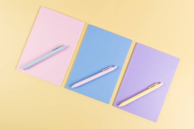 Sarı arka planda pembe, mor ve mavi defterlerin üst görüntüsü. Pembe, sarı ve mavi kalemler, okul, ofis kağıdı. Düz yat, boşluğu kopyala.