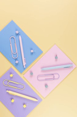 Sarı arka planda pembe, mor ve mavi defterlerin üst görüntüsü. Renkli kalemler, ataçlar, raptiyeler, okul, ofis kağıdı. Düz yat, boşluğu kopyala.