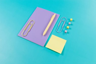 Mavi arkaplanda mor defterin üst görünümü. Yapışkan not, ataç, kalem ve iğne. Okul, ofis duvar kağıdı. Düz yat, boşluğu kopyala.