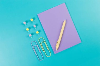 Mavi arkaplanda mor defterin üst görünümü. Ataç, kalem ve raptiye. Okul, ofis duvar kağıdı. Düz yat, boşluğu kopyala.