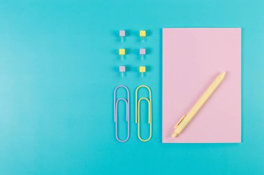 Mavi arkaplanda pembe defterin üst görüntüsü. Ataç, kalem ve raptiye. Okul, ofis duvar kağıdı. Düz yat, boşluğu kopyala.