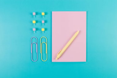 Mavi arkaplanda pembe defterin üst görüntüsü. Ataç, kalem ve raptiye. Okul, ofis duvar kağıdı. Düz yat, boşluğu kopyala.