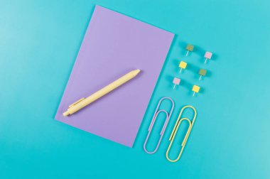 Mavi arkaplanda mor defterin üst görünümü. Ataç, kalem ve raptiye. Okul, ofis duvar kağıdı. Düz yat, boşluğu kopyala.