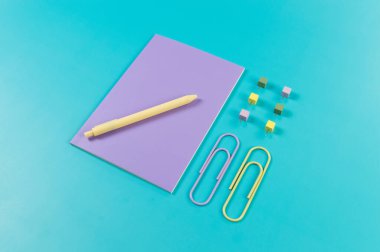 Mavi arkaplanda mor defterin üst görünümü. Ataç, kalem ve raptiye. Okul, ofis duvar kağıdı. Düz yat, boşluğu kopyala.