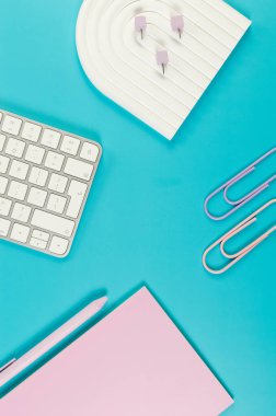 Beyaz klavyenin üst görüntüsü, pembe defter, mavi arka planda pembe kalem. Raptiyeler, raptiyeler, modern ofis, okul. Beyaz kemer tepsisi, düz konum, kopyalama alanı.