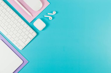 Beyaz klavye, fare, dokunmatik ped, kulaklık kutusu, mavi arka planda kalem. Mor ve pembe defterler, modern ofis, okul. Düz yat, boşluğu kopyala.