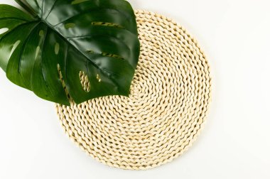 Kahverengi hasır masa paspası, beyaz arka planda yeşil canavar yaprağı. Saman yuvarlak tepsi, düz konum, kopyalama alanı. 