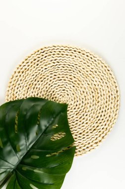 Kahverengi hasır masa paspası, beyaz arka planda yeşil canavar yaprağı. Saman yuvarlak tepsi, düz konum, kopyalama alanı. 