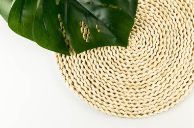 Kahverengi hasır masa paspası, beyaz arka planda yeşil canavar yaprağı. Saman yuvarlak tepsi, düz konum, kopyalama alanı. 