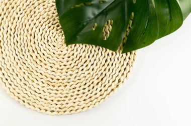 Kahverengi hasır masa paspası, beyaz arka planda yeşil canavar yaprağı. Saman yuvarlak tepsi, düz konum, kopyalama alanı. 
