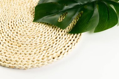 Kahverengi hasır masa paspası, beyaz arka planda yeşil canavar yaprağı. Saman yuvarlak tepsi, düz konum, kopyalama alanı. 