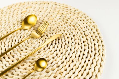Kahverengi hasır masa paspası, beyaz arka planda altın çatal bıçak takımı. Saman yuvarlak tepsi, düz konum, kopyalama alanı. 