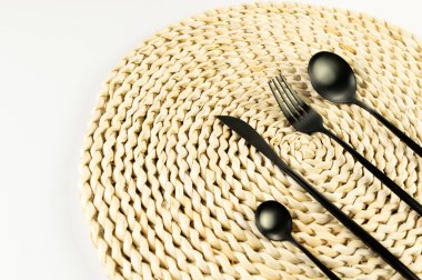 Kahverengi hasır masa paspası, beyaz arka planda siyah çatal bıçak takımı. Saman yuvarlak tepsi, düz konum, kopyalama alanı. 