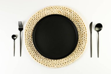 Kahverengi hasır masa paspası, siyah tabak, beyaz arka planda siyah çatal bıçak. Saman yuvarlak tepsi, düz konum, kopyalama alanı. 