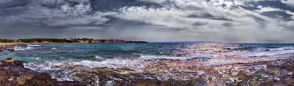 Dramatic sky and bad weather panoramic view at wild rocky bay  over sea with waves crashing at the shore with overcast rainy sky and sunbeams hits water in a suggestive and impressive seascape.