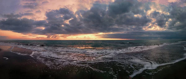Panoramic cloudy sunset at the beach with dramatic sky and dark stratocumulus clouds with orange sunlight, dark sand with rough sea and lots of white sea foam.