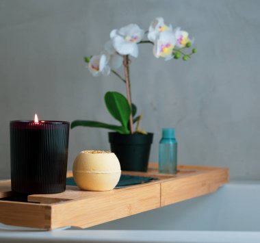 Ahşap bir banyo tepsisi, yanan bir mum, güzel kokulu bir banyo bombası ve zarif beyaz orkidelerle dolu bir vazo..
