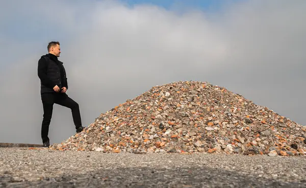 Bir adam dramatik bulutlu gökyüzünün altında muazzam bir çakıl yığınının önünde güvenle poz verir.