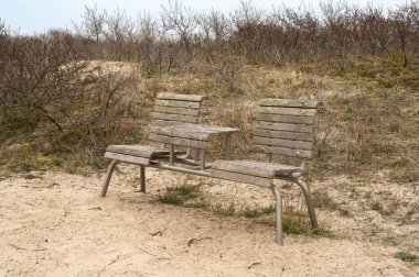 Eski, yıpranmış ahşap bir bank kumlu zeminde uzanır. Etrafı seyrek ve az bitki örtüsüyle çevrilidir.