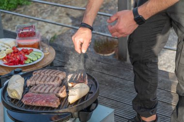 Bir şef, taze sebzeler ve sakin bir açık hava ortamı ile çevrili ızgaradaki bifteği ustalıkla pişirir..