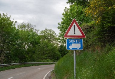 Yeşilliklerle çevrili virajlı bir yolda yaklaşan bir araç çıkışını gösteren bir uyarı levhası..