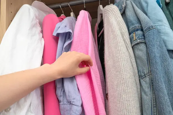 Bir insan, dolapta asılı duran canlı pembe bir kazağa uzanmak için kolunu uzatıyor ve rahat bir kıyafet arıyor.