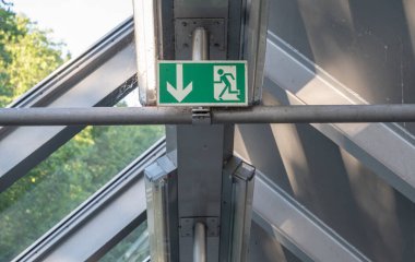 Aşağıya doğru bir kaçış rotası belirten bir acil çıkış levhası, modern cam bir binada yer alıyor.