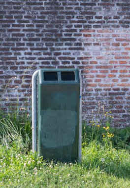 Yeşil bir çöp kutusu kırsal tuğla bir duvara karşı duruyor, çimenler ve renkli kır çiçekleriyle çevrili.