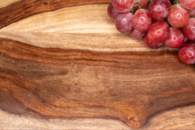 Tahta kesme tahtasının kenarında duran bir grup kırmızı üzüm negatif boşluk bırakıyor. Kırsal ahşap dokusu canlı meyveyi tamamlıyor, yemek fotoğrafçılığı için ideal..