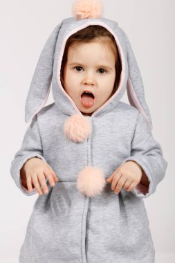 Yumuşak pastel tavşan kostümü giymiş, yumuşak kulaklı ve pofuduk ponponlu şirin bir bebek komik bir ifade takınıyor, dillerini çıkarıp, şakacı bir şekilde ellerini kaldırıyor. Paskalya için mükemmel