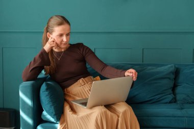 Thoughtful woman sitting on a teal sofa with a laptop on her lap. Concentrated and focused expression. Casual home office setup.