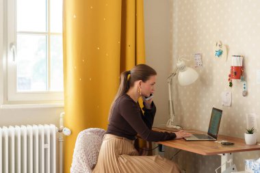 Cheerful woman working remotely from her home office, sitting at a desk with a laptop and talking on the phone. The bright interior, yellow curtain, and casual outfit convey a modern work-from-home lifestyle.
