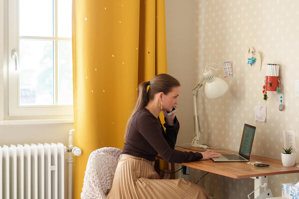 Cheerful woman working remotely from her home office, sitting at a desk with a laptop and talking on the phone. The bright interior, yellow curtain, and casual outfit convey a modern work-from-home lifestyle.