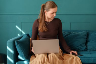 Woman sitting on a teal couch, working on her laptop while holding a smartphone. She is dressed casually in a brown top and beige skirt. Bright and modern interior.