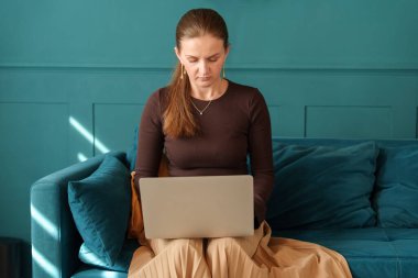 Woman sitting on a teal sofa, focused on her laptop. She is casually dressed and working in a cozy home environment. Concept of remote work or studying.