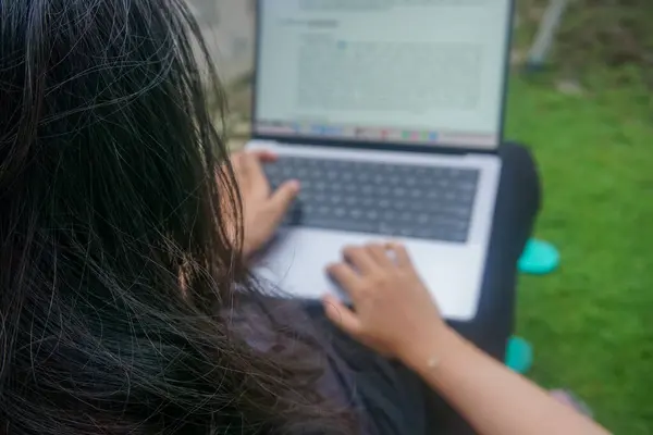 Bokeh. Öğrenci bir kız, öğleden sonra bahçede dizüstü bilgisayar kullanarak üniversite ödevini yazıyor. Bulanık bir arkaplan ile fotoğraflandı