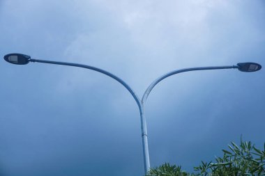 Modern bir sokak lambası direği. İki koluyla birlikte parlak mavi gökyüzüne yükseliyor. Arka planda beyaz bulutlar var.