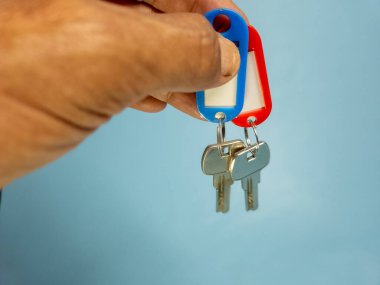 Bir adam açık mavi arka planda iki anahtar zinciri tutuyor.