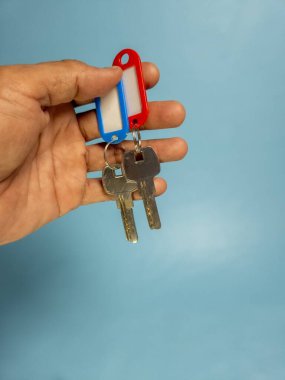 Bir adam açık mavi arka planda iki anahtar zinciri tutuyor.