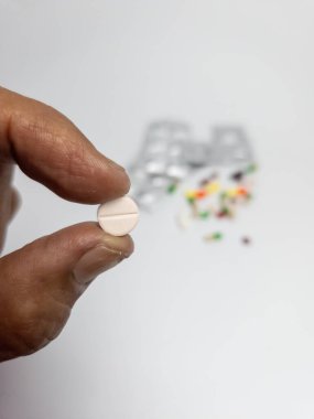 Yuvarlak, beyaz bir hap tutan, su toplayan ve arka planda gevşek haplar dağıtan bir elin yakın plan çekimi. Görüntü, ilaç, sağlık ve tedavi temalarını iletiyor. Sığ bir alan derinliği ile vurulmuş..