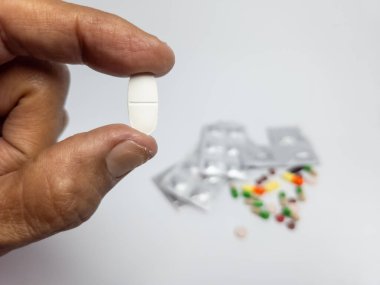 Arka planda su toplayan bir paket ve diğer haplarla birlikte beyaz bir hap tutan bir eli kapat. Sağlık ve tıbba odaklan. Beyaz yüzeyde doğal ışıkla çekilmiş..