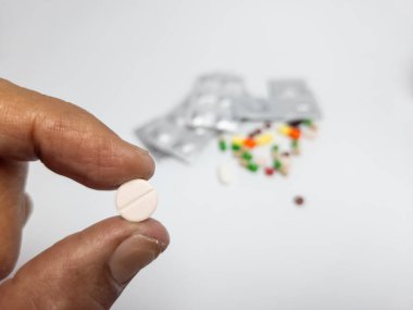 Beyaz yuvarlak hap tutan bir elin yakın çekimi. Bulanık arka planda su toplamaları ve renkli haplar var. İlaç, sağlık, tedavi ve ilaç kavramlarını temsil ediyor. Beyaz arkaplanda stüdyo görüntüsü.