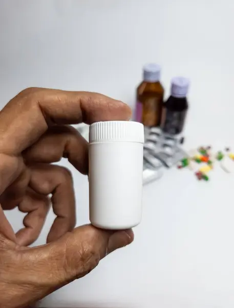 Bir el beyaz bir ilaç şişesini arka planda su toplamalı, renkli haplar ve ilaç şişeleriyle tutuyor. Bu görüntü sağlık, ilaç yönetimi ve ilaç endüstrisini temsil etmektedir..