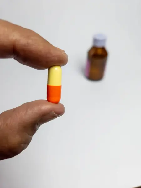 Arka planda küçük bir ilaç şişesi bulanık sarı ve turuncu bir kapsülü tutan bir elin yakın çekimi. Görüntü bir sağlık, ilaç ve tedavi duygusu taşır. İlaç ve sağlıkla ilgili projeler için uygundur.