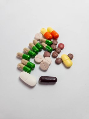 Yüksek açılı çekimde beyaz bir yüzeye dağılmış renkli haplar ve kapsüller bulunur. Düzenleme çeşitli şekiller, boyutlar ve renkler içererek, görsel olarak farklı bir ilaç koleksiyonu oluşturur..