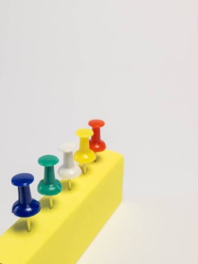 Sarı dikdörtgen bir bloğun beş renkli iğne sırasına dizilmiş yakın çekimi. İtme iğneleri sırasıyla mavi, yeşil, beyaz, sarı ve kırmızıdır. İğneler bloğa yerleştiriliyor ve düzgünce hizalanıyor. Arkaplan ne kadar sade