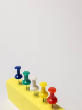 Sarı dikdörtgen bir nesnenin düz beyaz bir arkaplan üzerinde düzgün sıralanmış renkli itme iğnelerinin yakın çekim görüntüsü. Çiviler çeşitli renklerdedir: mavi, yeşil, beyaz, sarı ve kırmızı. Görüntü iyi aydınlatılmış ve keskin odaklı, vurgulu.