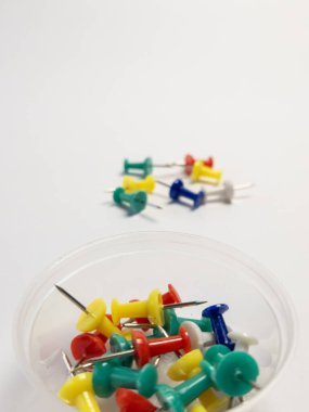 Renkli raptiye koleksiyonu sergileyen yakın plan bir fotoğraf. Bazıları arka planda dağılırken, diğerleri ön plandaki küçük temiz plastik bir kabın içine yerleştiriliyor. Görüntü beyaz bir arkaplanda çekiliyor ve canlı koloyu vurguluyor