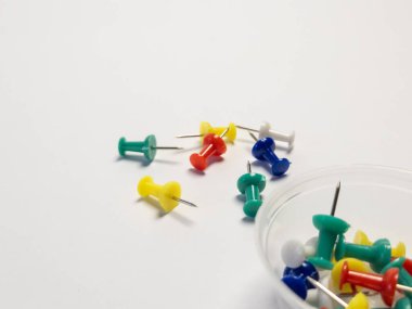 Yeşil, sarı, kırmızı ve maviyi içeren renkli bir itme iğnesi koleksiyonu düz beyaz bir yüzeye saçılmıştır. Bazı raptiyeler de temiz bir plastik kutunun içinde. Görüntü aydınlık, temiz ve düzenli bir his yaratıyor, mükemmel. 